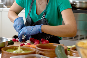 woman cutting radishes on wood, sitting at kitchen table. green t-shirt and denim apron, blue vinyl gloves.