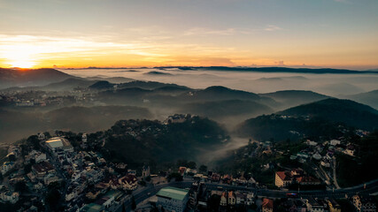Aerial view of Dalat City at dawn, showcasing mist-covered mountains, lush greenery, scattered...