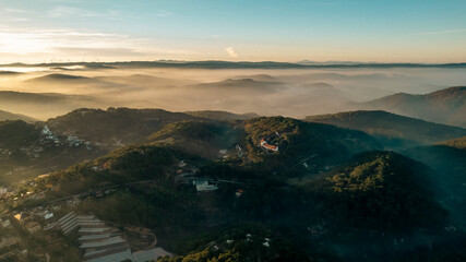 Aerial view of Dalat City at dawn, showcasing mist-covered mountains, lush greenery, scattered...