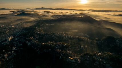 Aerial view of Da Lat at sunrise, showcasing mist-covered buildings amidst lush mountains, under a golden sky, highlighting the city’s architectural and natural beauty.