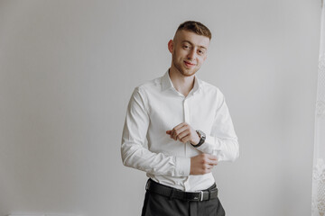 A man in a white shirt and black pants is standing in front of a white wall. He is wearing a watch and is adjusting his shirt. Concept of formality and attention to detail