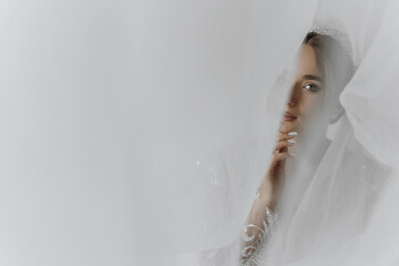 A woman is standing in front of a curtain, looking at the camera. She is wearing a white dress and...