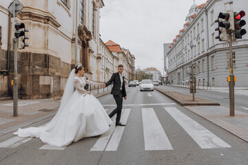 A bride and groom are walking across a street. The bride is wearing a white dress and the groom is wearing a suit. The couple is holding hands and walking together