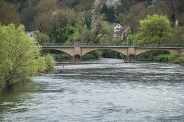 Fototapeta premium Rundbogenbrücke aus Sandstein mit halbrunden Strömungsteilern vor Bäumen mit frischem Blattgrün und versteckten Häusern an Fluss Neckar mit silbern glänzenden Reflexionen bei Neckartenzlingen. 