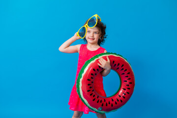 happy little girl in sunglasses holds a swimming circle in the shape of a watermelon on a blue...