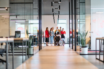 A diverse group of young business people walking a corridor in the glass-enclosed office of a modern startup, including a person in a wheelchair and a woman wearing a hijab