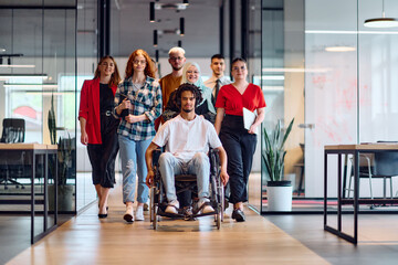 A diverse group of young business people walking a corridor in the glass-enclosed office of a modern startup, including a person in a wheelchair and a woman wearing a hijab