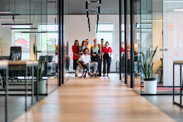 A diverse group of young business people walking a corridor in the glass-enclosed office of a modern startup, including a person in a wheelchair and a woman wearing a hijab