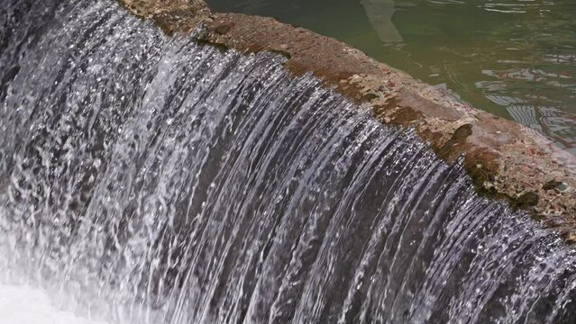 water flowing over concrete barrier in a row of thin trickles in small river dam at sunny day, closeup with slow motion
