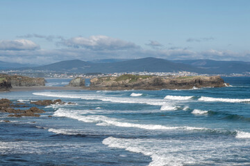 Scenic coastline, rocky shores, waves crashing, clear sky, distant mountains, peaceful exploration, untouched environment.