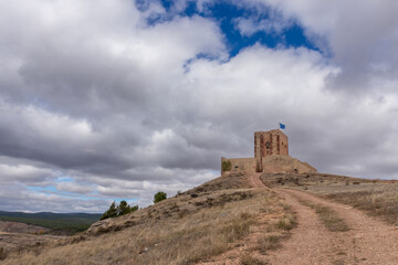 An old stone structure on a hill, path leading up, flag on top, surrounded by a rugged, cloud-shadowed landscape