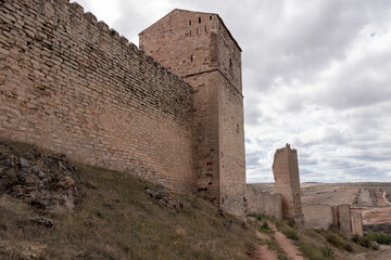 an ancient, tall stone wall and tower, part of a fortress, under a cloudy sky with barren...