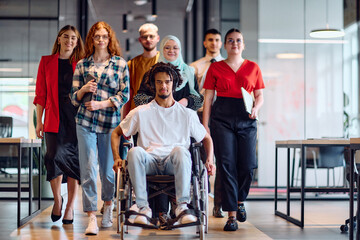 A diverse group of young business people walking a corridor in the glass-enclosed office of a modern startup, including a person in a wheelchair and a woman wearing a hijab