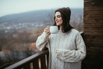 Young woman in white knitted sweater holding white cup of tea, standing on a balcony of a wooden house with mountains view.