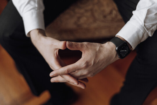 A man is sitting on a chair with his hands folded in his lap. He is wearing a watch and a white shirt. Concept of calmness and relaxation - Powered by Adobe