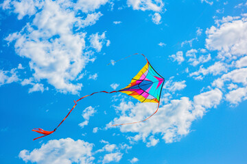 A captivating shot of a colorful kite soaring high above the ground, against a backdrop of clear blue sky and fluffy white clouds.