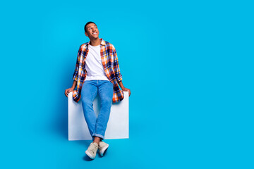 Full size photo of young man sit cube look empty space wear shirt isolated on blue color background