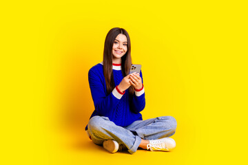 Full length photo of pretty teen girl hold device crossed legs wear trendy knitwear blue outfit isolated on yellow color background