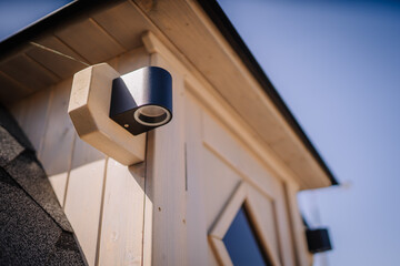 A close-up view of a modern outdoor lighting fixture mounted on the wooden wall of a tiny house, with blurred trees in the background.