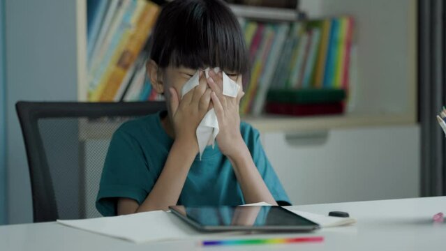 An Asian kid girl, age 8+, does her homework on a tablet in her home office. Her nose felt stuffy, and she quickly grabbed a tissue and sneezed and wiped her nose well. The background is a bookshelf