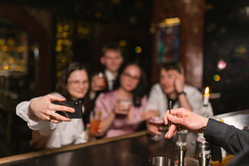 Happy guests group makes selfie and bartender makes beverage at counter focus on hands. Fancy...