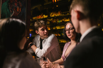 Girl invites friends spend time together at weekend at club. Guys talk with girls with glasses and drink alcoholic cocktails in evening