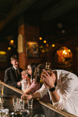 Depressive man holds head by hand on bar counter. Guy with alcohol intoxication sits after noisy...