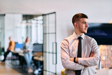 A young business leader stands with crossed arms in a modern office hallway, radiating confidence...