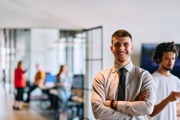 A young business leader stands with crossed arms in a modern office hallway, radiating confidence...