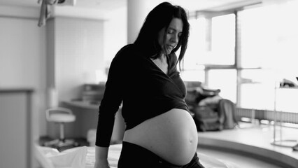 Dramatic scene of pregnant woman seated by bedside at hospital having contractions struggling with pain during pre labor, breathing deeply suffering during childbirth in monochrome