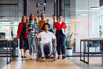 A diverse group of young business people walking a corridor in the glass-enclosed office of a modern startup, including a person in a wheelchair and a woman wearing a hijab