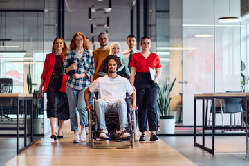 Obraz premium A diverse group of young business people walking a corridor in the glass-enclosed office of a modern startup, including a person in a wheelchair and a woman wearing a hijab