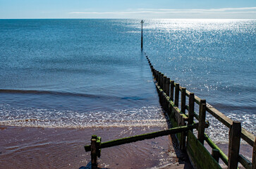 Beach sand groyne leading out to the ocean, water on a sunny day. blue sky. Calm, reflective and...