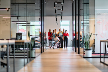 A diverse group of young business people walking a corridor in the glass-enclosed office of a modern startup, including a person in a wheelchair and a woman wearing a hijab