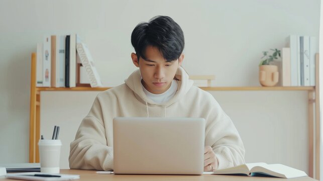 Focused young Asian man studying or working on a laptop in a neatly organized workspace with books and a cup.