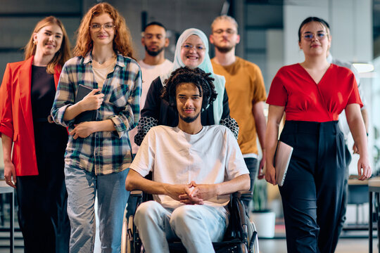 A diverse group of young business people walking a corridor in the glass-enclosed office of a modern startup, including a person in a wheelchair and a woman wearing a hijab