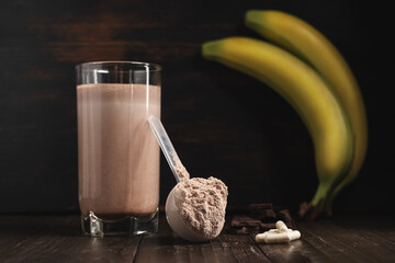 Plastic measuring spoon with whey protein powder, milkshake cocktail in a glass, blended protein drink, white pills or capsules, chocolate cubes and banana fruit on a dark wooden background