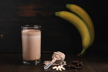Plastic measuring spoon with whey protein powder, milkshake cocktail in a glass, blended protein drink, white pills or capsules, chocolate cubes and banana fruit on a dark wooden background