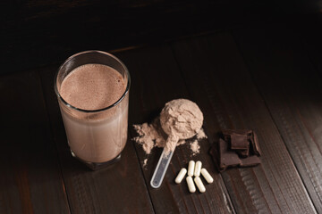 Protein milkshake cocktail in a glass, plastic measuring spoon with whey protein powder, white pills or capsules of amino acids, chocolate cubes on a dark wooden background