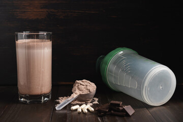 Protein milkshake cocktail in a glass, plastic measuring spoon with whey protein powder, white pills or capsules of amino acids, chocolate cubes on a dark wooden background