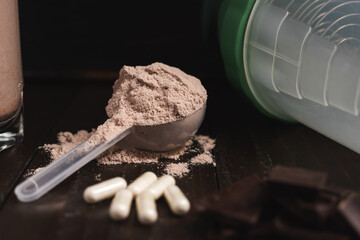 Plastic measuring spoon with whey protein powder, milkshake cocktail in a glass, blended protein drink, white pills or capsules, chocolate cubes on a dark wooden background