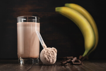 Plastic measuring spoon with whey protein powder, milkshake cocktail in a glass, blended protein drink, chocolate cubes and banana fruit on a dark wooden background