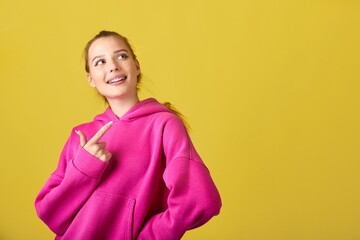 A joyful young woman with braces on her teeth in a stylish pink hoodie smiling and pointing to the...