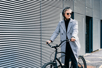Stylish male in coat, sunglasses and protective helmet riding on retro bicycle near gray urban...
