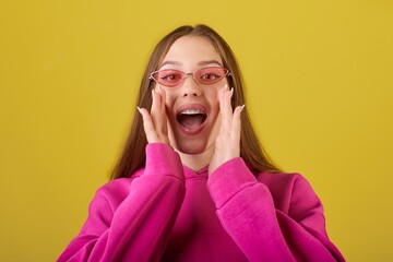 Young woman in a vibrant pink hoodie and stylish glasses, energetically expressing excitement with hands by her face, shouting joyfully with an open mouth against a cheerful yellow background.