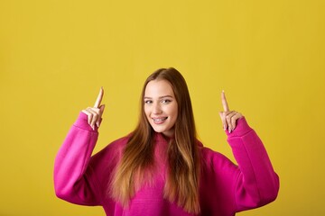 A young woman in a pink pullover points upwards with a happy expression on her face. She has long...