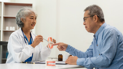 Senior Asian female doctor showing a dental prosthetic model to a patient with toothache discussing medical treatment during a dental consultation in the office.