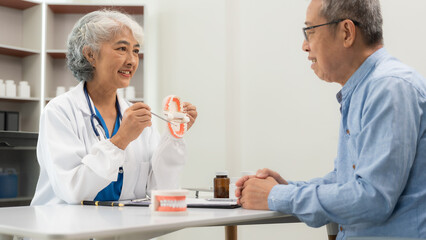 Senior Asian female doctor showing a dental prosthetic model to a patient with toothache discussing...