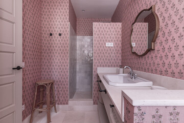 modern design bathroom with cream marble sink, golden wood framed mirror, matching furniture and walls with decorative pink wallpaper and natural cane stool