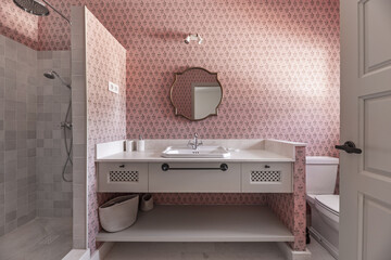 A beautiful modern designed bathroom with cream marble sink, gold wooden framed mirror, matching furniture and walls with decorative pink wallpaper and a shower cubicle with tiled walls